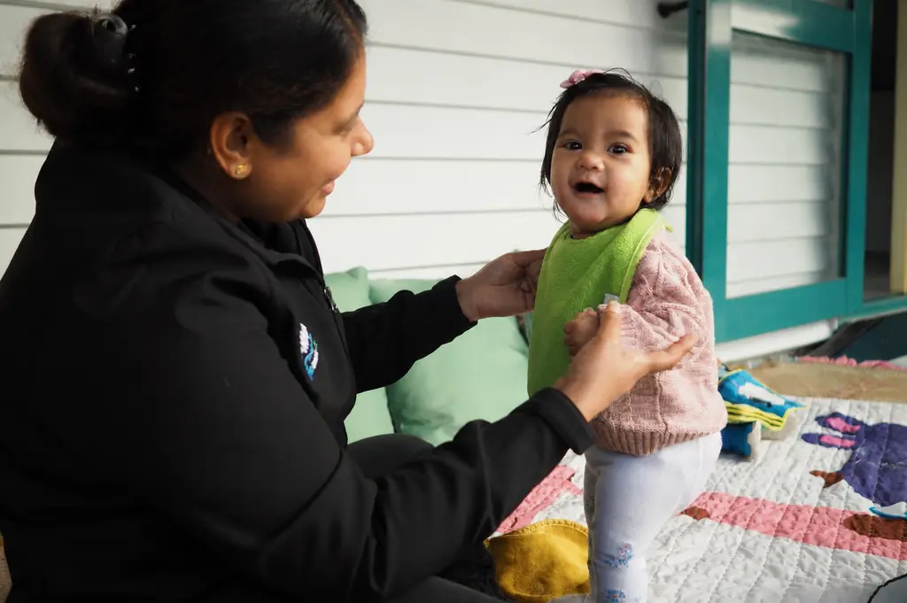 Smiling infant supported by a kaiako during outdoor play