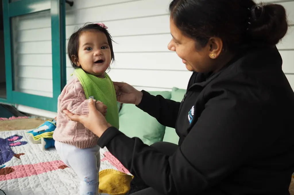 Kaiako supporting a smiling infant during outdoor play